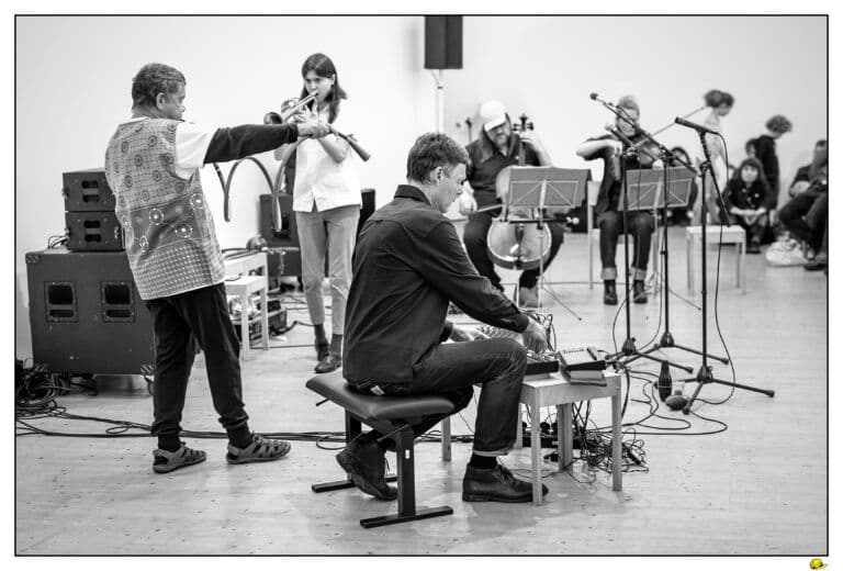 Black-and-white photo of musicians rehearsing: a man holds out an electrified bow, a woman plays a wind instrument, another man sits at electronic gear, with cellist and violinist behind.