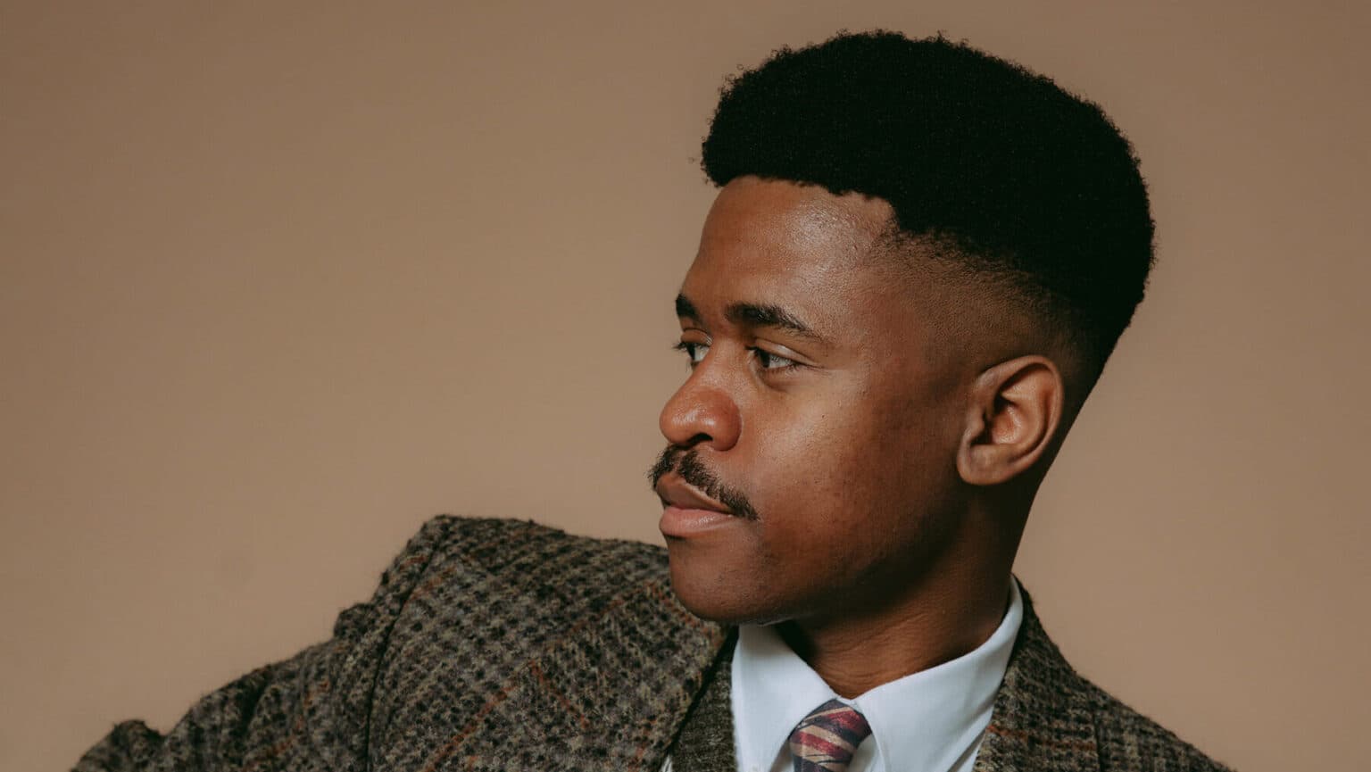 Portrait of singer Booshle G in profile, wearing a tweed jacket and striped tie against a warm beige backdrop.