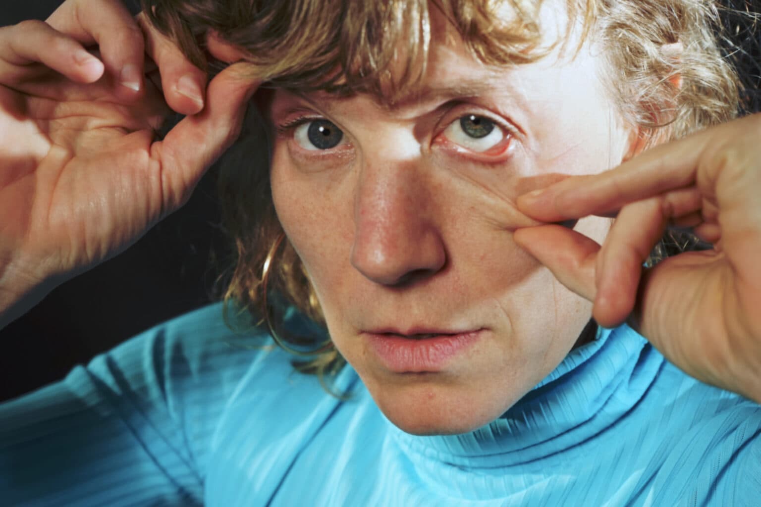 Close-up portrait of the singer in a bright blue turtleneck, hands raised to their face, looking straight at the camera.