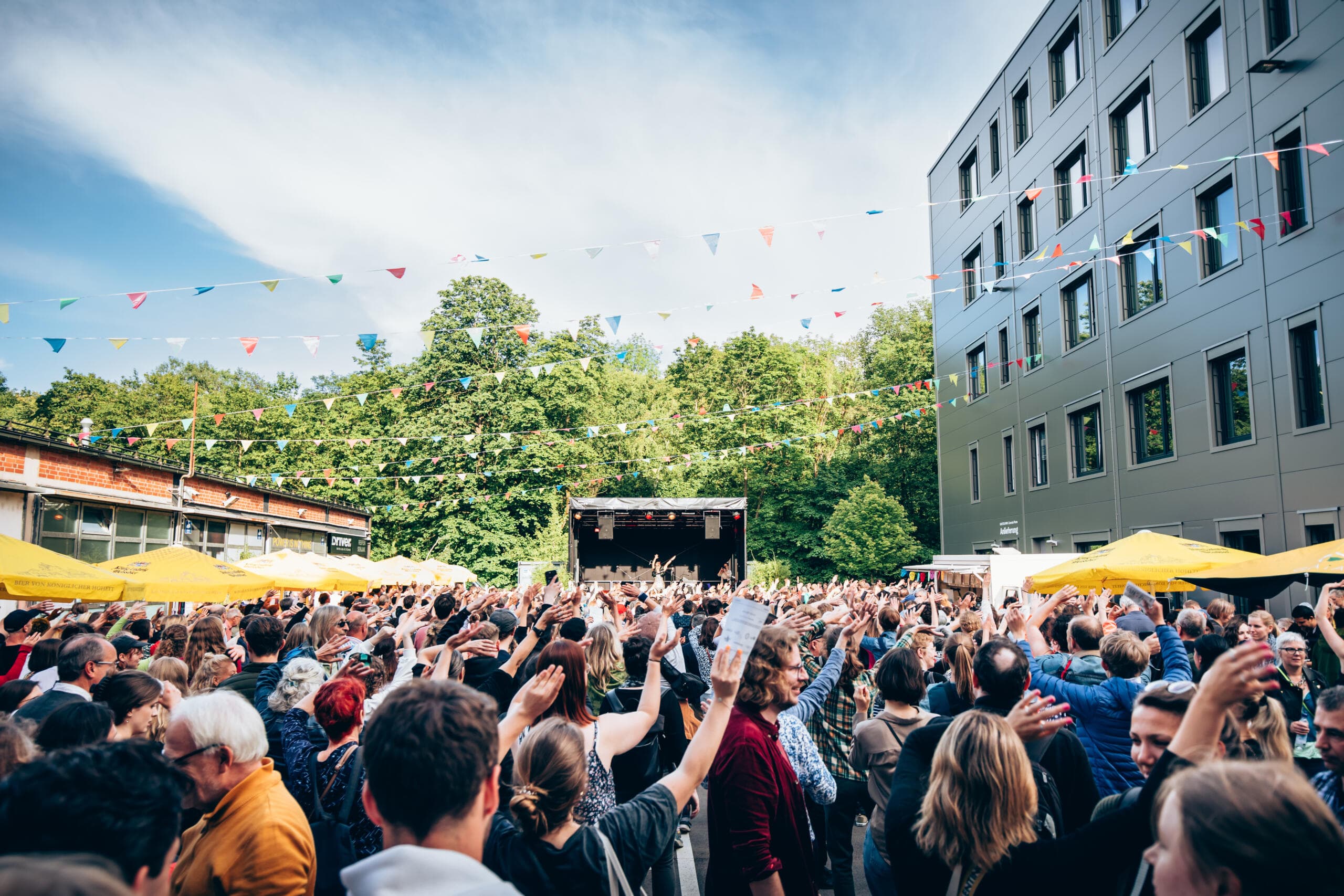 A large crowd with arms raised gathers under colourful bunting on the open square outside Gasteig HP8, cheering towards an outdoor stage on a sunny day.