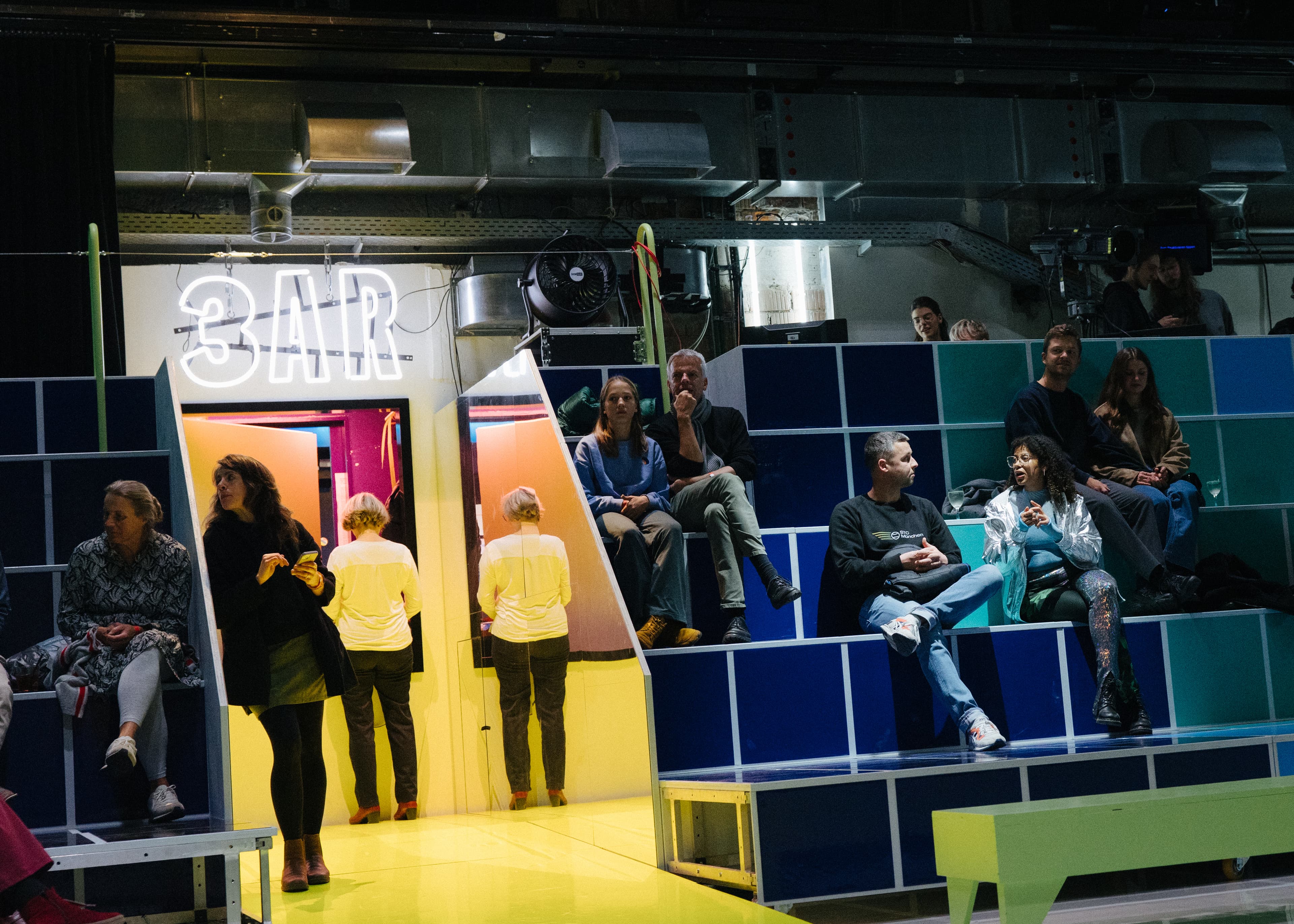 Guests sitting and standing on colourful blue and yellow tiled bleacher seating in the Werkraum, with a glowing neon sign and dressing-room mirrors on a bright yellow floor.