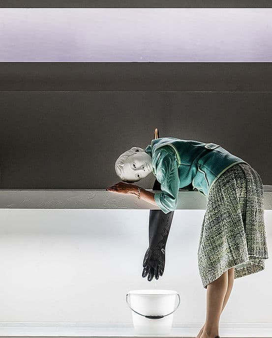 Staged scene from La Musica: a pale-faced figure in a green jacket and tweed skirt drapes over a white ledge, one arm in a long black glove pointing down towards a white bowl of water.