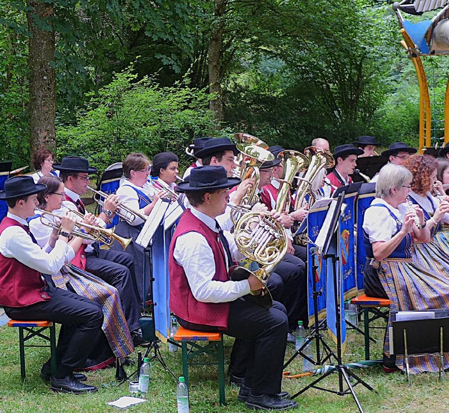 The Blaskapelle Unterföhring performs outdoors among trees in red waistcoats and traditional dress.