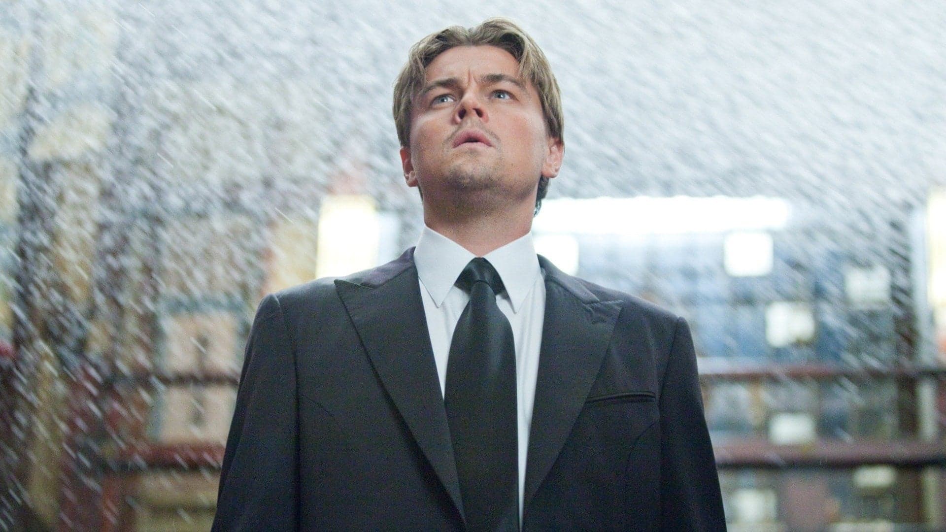 Young person in a dark suit and tie stands centered, looking upward with mouth slightly open as fine white streaks fall around it; low-angle, backlit.