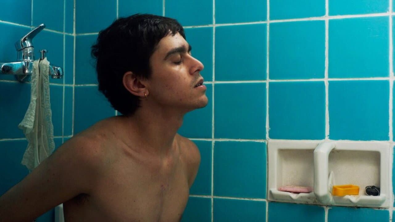 Young shirtless person with short dark hair and a small hoop earring leans back in a narrow shower, eyes closed and head tilted; turquoise tiled wall, faucet draped with lace, soap niche with bars.