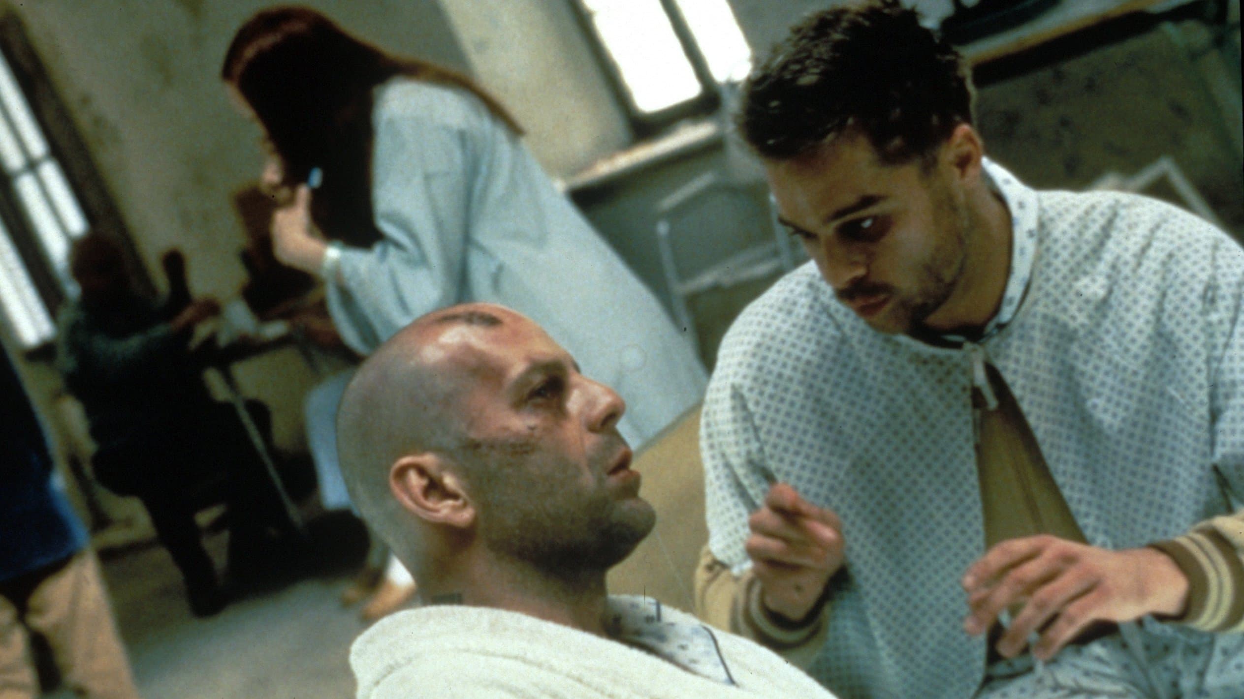 A hospital-gowned person leans toward a seated bald person with a forehead bruise, speaking and gesturing in a dim ward lit by cool side light.