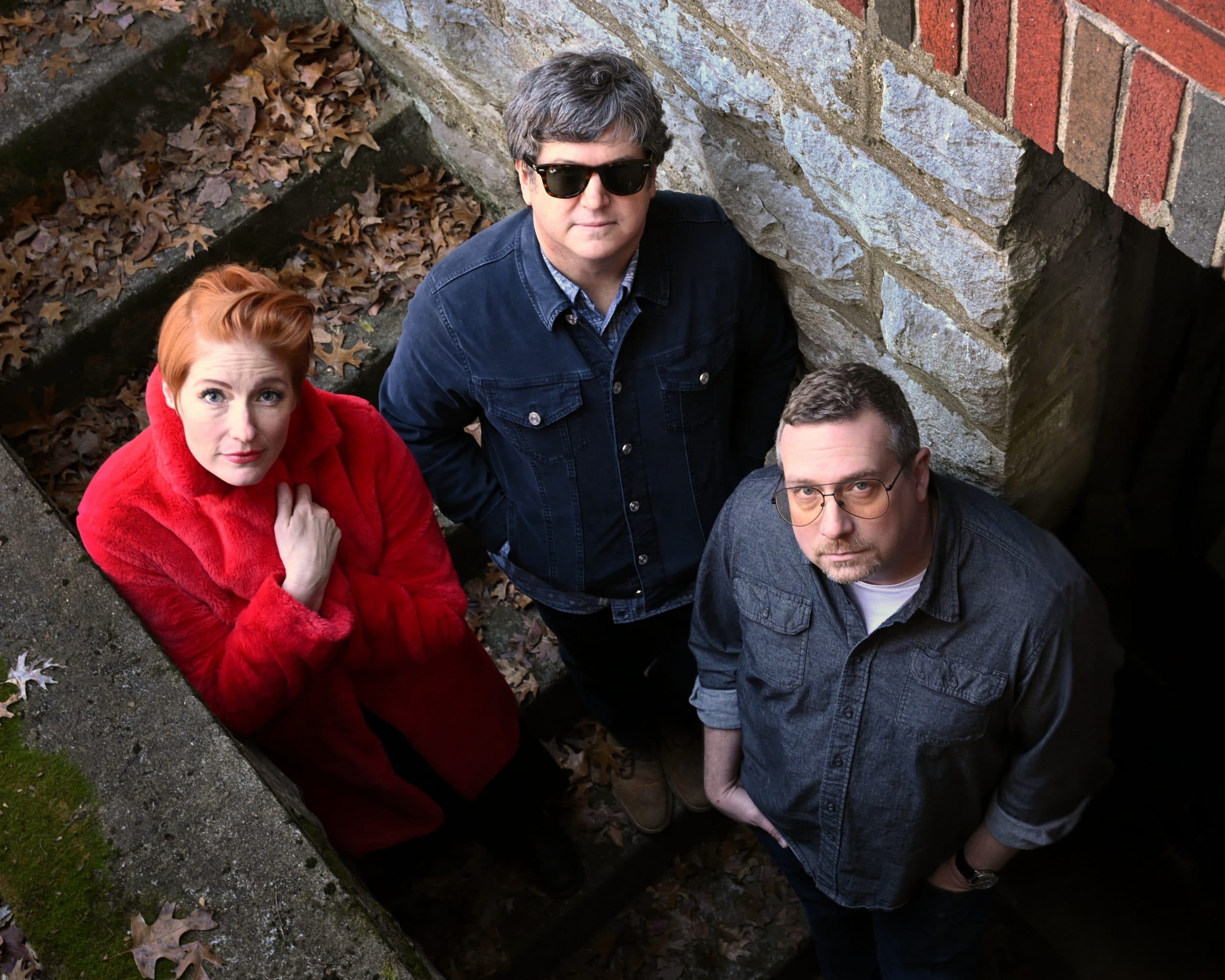 The three members of Sixpence None The Richer photographed from above in a leaf-strewn stairwell, one wearing a red coat.