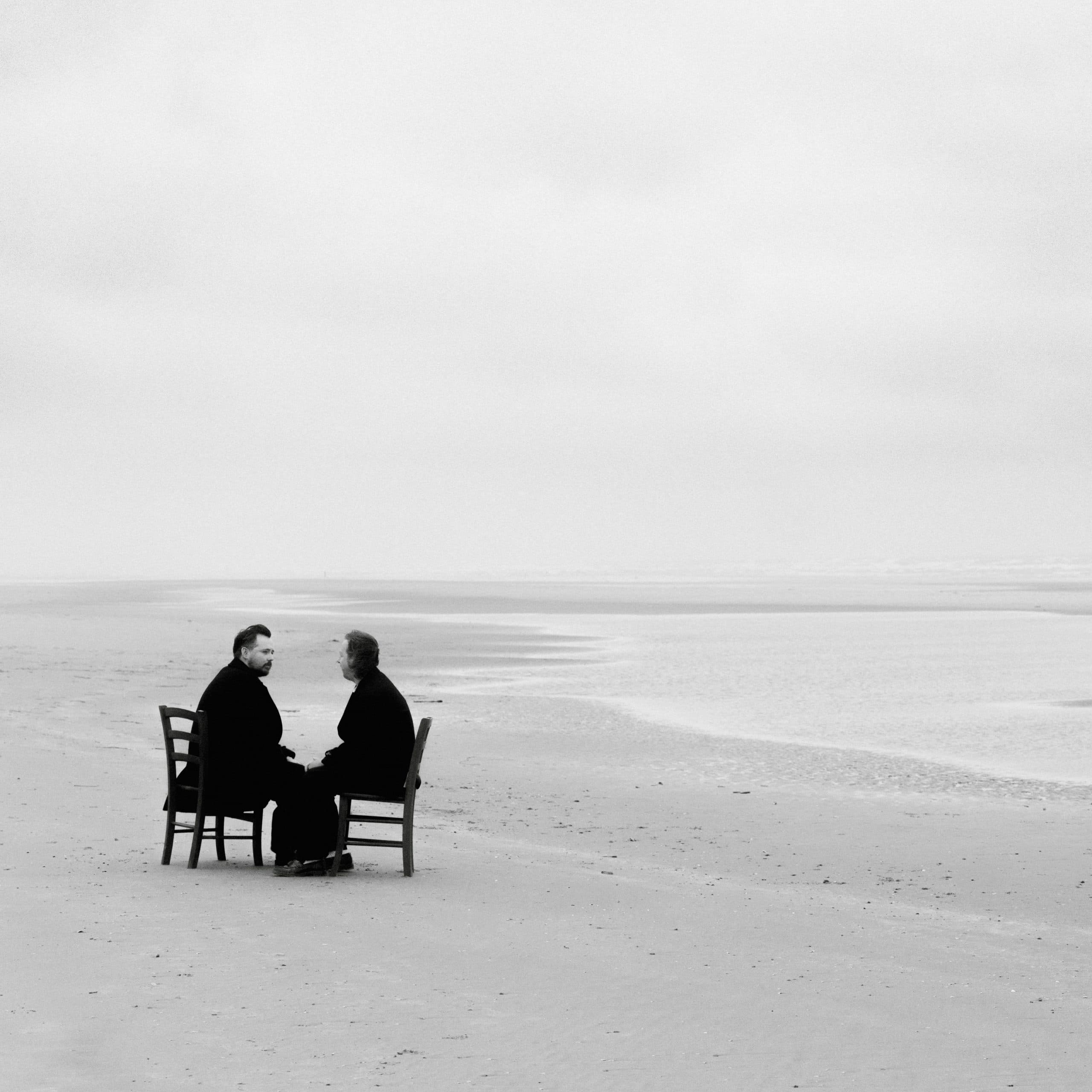 Black-and-white press photo of the two members of Tender sitting on wooden chairs facing each other on a wide, empty beach.