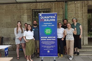 Eine Gruppe von acht Personen steht zusammen vor einem blauen Roll-up-Banner mit der Aufschrift "QUANTEN(T)RÄUME" und Projektlaufzeit 1.12.2023–30.11.2026; Banner bewirbt "KEINE WOCHE OHNE QUANTEN!" und zeigt ein grünes Stern-Schild mit Hinweis auf wöchentlichen Quantendienstag. Szene vor einer steinernen Gebäudefassade mit Fenster und grünem Rohr; einige Personen tragen Freizeitkleidung, links ein Tisch mit Flyern. Gesichter absichtlich unkenntlich gemacht.