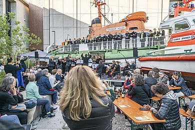 Publikum sitzt an Bierzeltgarnituren und auf Stufen, während ein Orchester und ein Chor auf der Rampe vor zwei Rettungsbooten (Schiffe beschriftet "THEODOR HEUSS" und "ASMUS BREMER") musizieren; Dirigent hebt die Arme, viele Zuschauer fotografieren mit Smartphones.