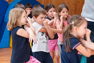 Eine Gruppe von Grundschulkindern in Turnhalle macht Atemübungen: mehrere Mädchen und ein Junge stehen dicht beieinander, halten sich die Nasen oder pressen Finger an die Nasenwurzel, tragen bunte Freizeitkleidung (T-Shirt, Shorts), im Hintergrund blaue Weichbodenmatten und Holzverkleidung.