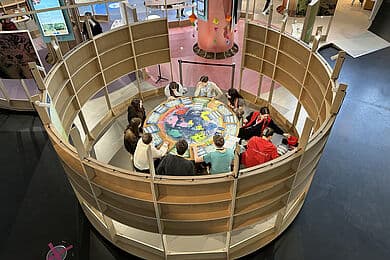 Zehn Menschen sitzen an einem runden Tisch, auf dem eine große Weltkarte abgebildet ist und spielen ein Spiel.