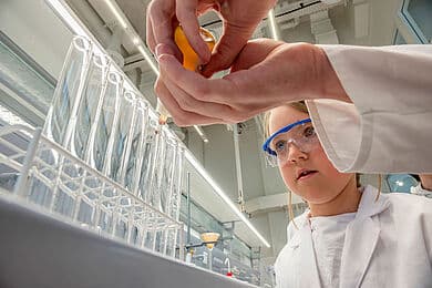 Ein Kind in einem weißen Laborkittel und blauer Schutzbrille beobachtet aufmerksam ein chemisches Experiment im Chemielabor. Eine andere Person, deren Hände sichtbar sind, pipettiert eine orangefarbene Flüssigkeit in ein Reagenzglas, das sich in einem Gestell mit mehreren leeren Reagenzgläsern befindet.