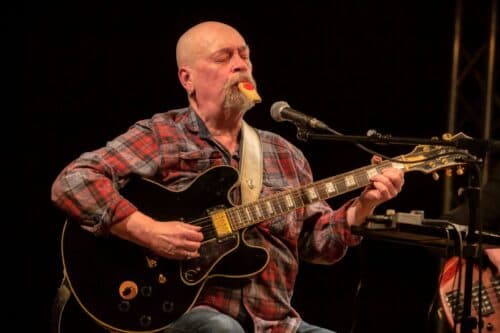 Schorsch Hampel sings into a microphone while playing electric guitar onstage.