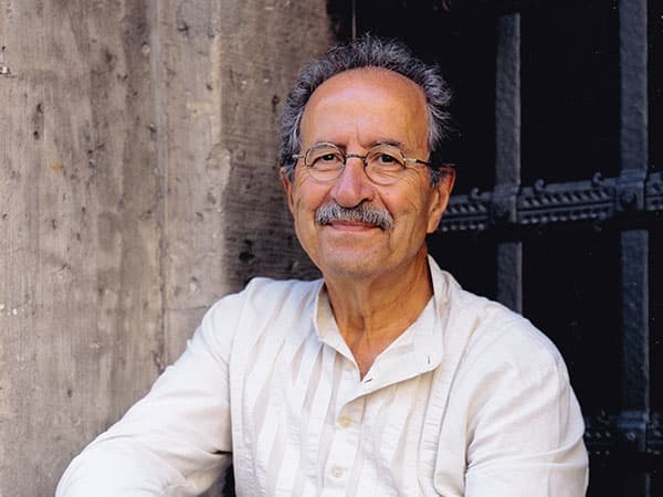 Portrait of author Rafik Schami, an older man with grey hair, a grey moustache and glasses, smiling in a pale open-necked shirt against a weathered wooden door.