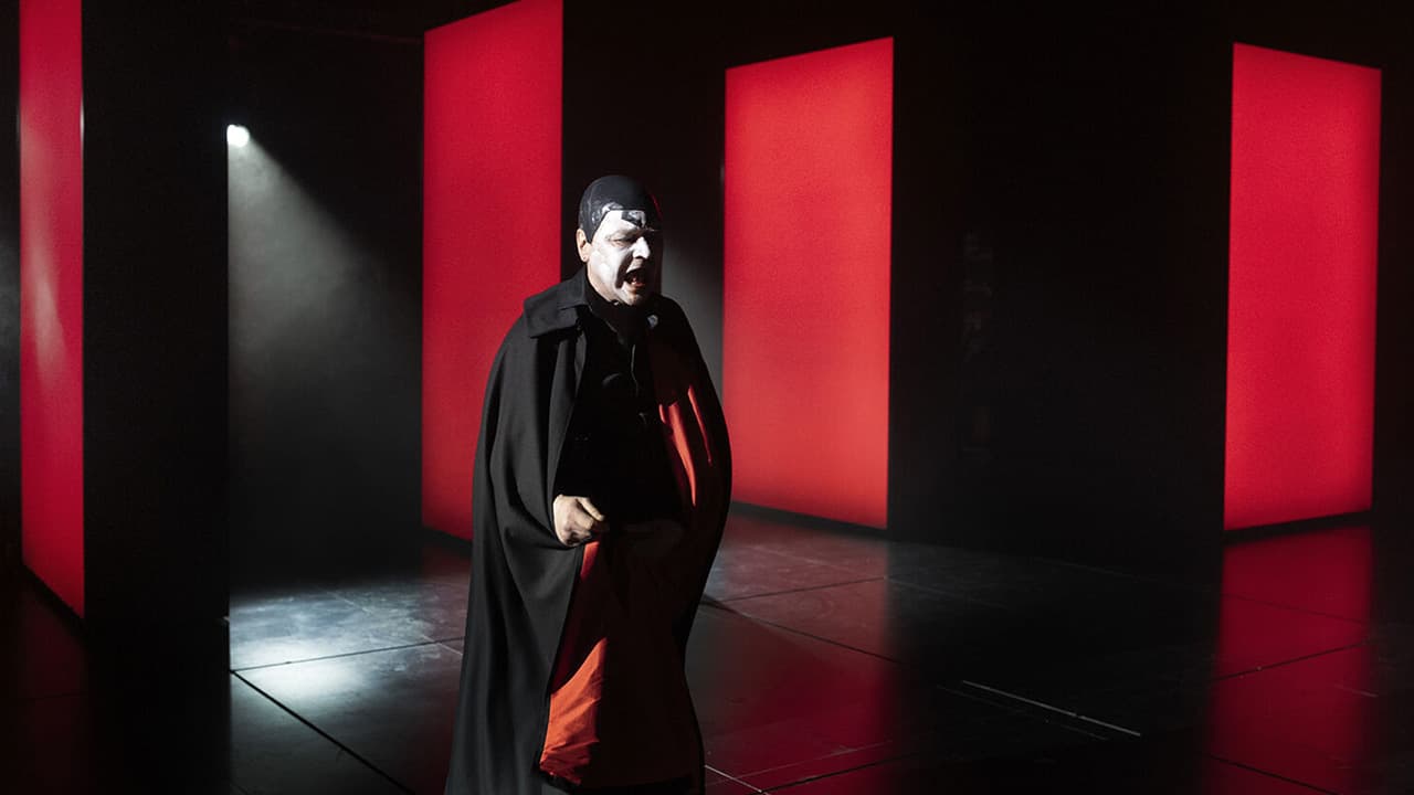 An actor in white and black stage makeup and a long black cape stands on a dark stage lined with tall glowing red panels.