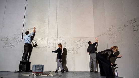 Four performers stand at a huge pale wall covered in handwritten names, each writing or reaching up with chalk in a bare theatre space.